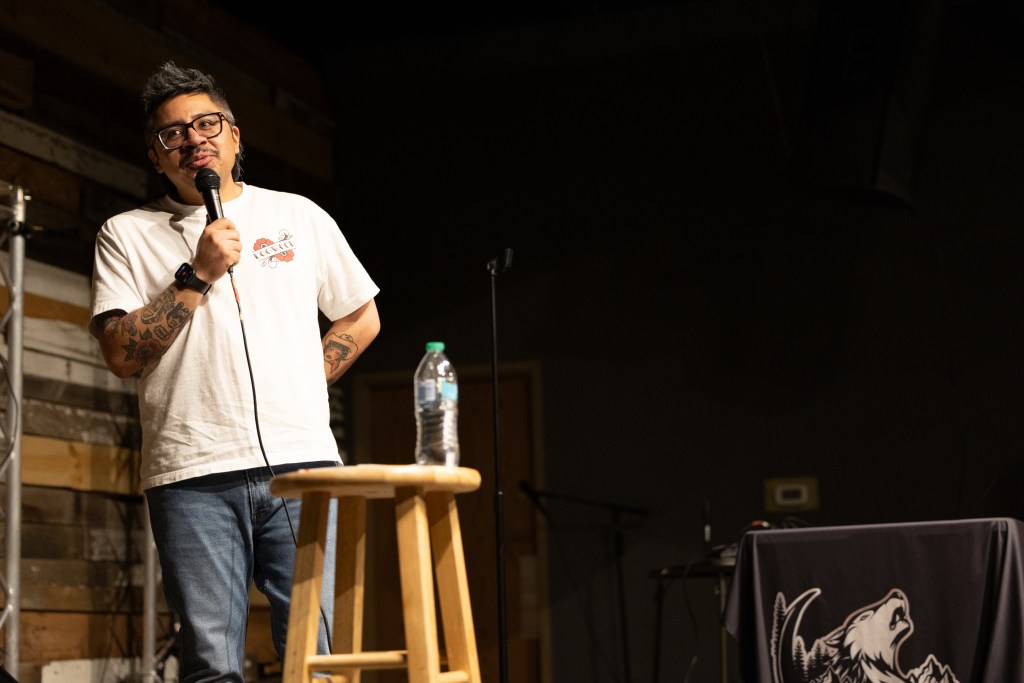 A comedian performing on stage with a microphone, wearing a white t-shirt and jeans, standing next to a wooden stool with a water bottle on it.
